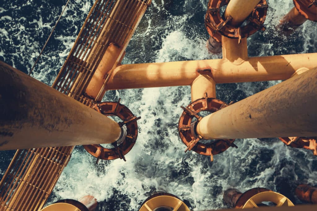 Overhead view of yellow offshore platform pipes and metal walkways above churning ocean water.